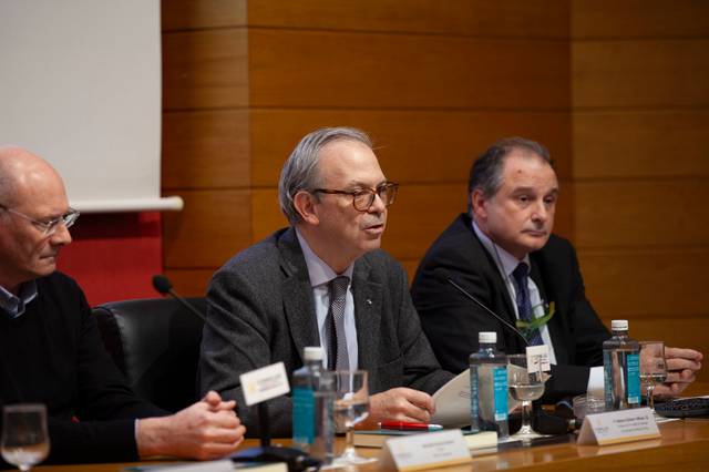 Tres hombres sentados en una mesa durante una conferencia, hablando ante micrófonos y con botellas de agua delante de ellos.