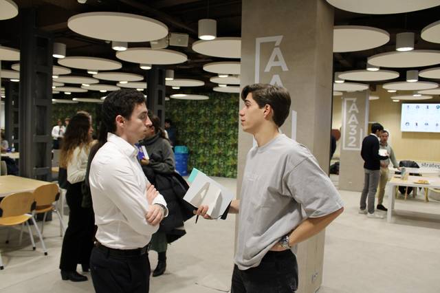 Dos hombres conversan en un ambiente laboral moderno con otras personas al fondo.