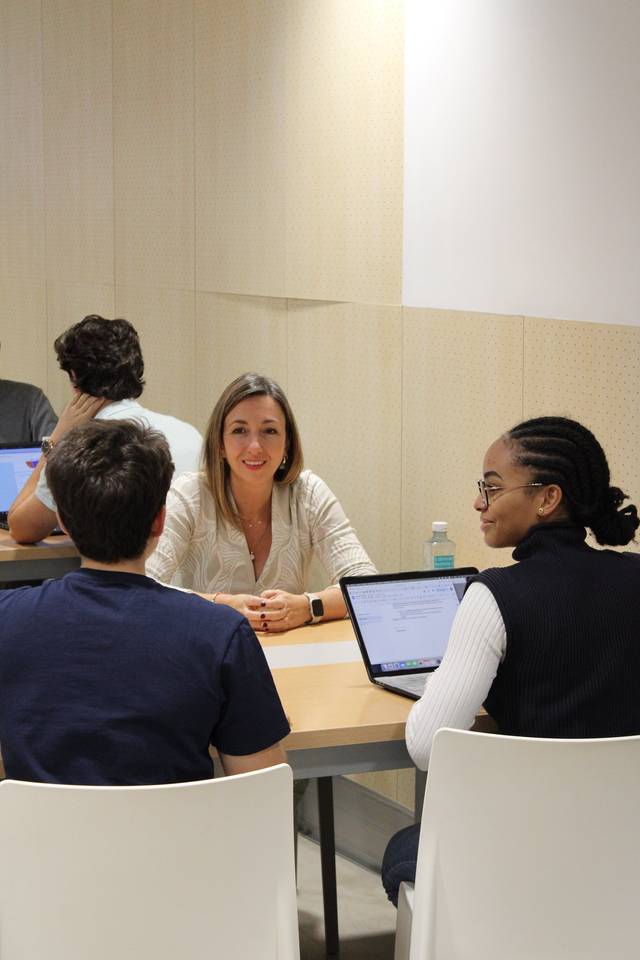 Tres personas sentadas alrededor de una mesa en una sala de trabajo colaborativo, dos de ellas conversando mientras usan sus computadoras portátiles.