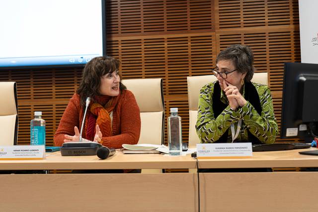 Dos mujeres conversando en un panel de conferencia, sentadas frente a micrófonos y botellas de agua, con placas de identificación visibles.