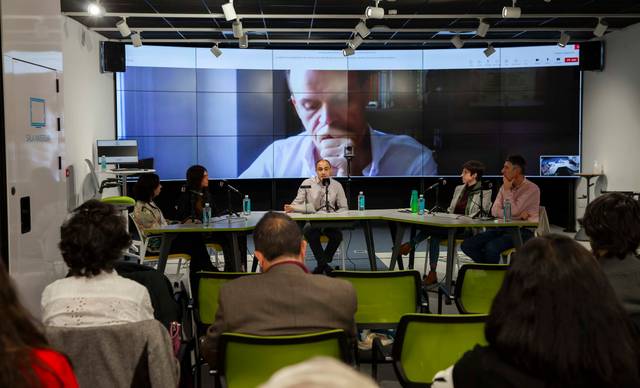 Personas sentadas en una sala de conferencias mientras observan a un hombre hablando en una pantalla grande.