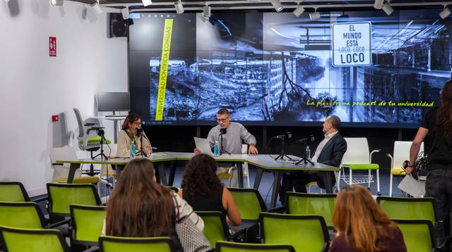 Panelistas discutiendo en un evento en una sala con sillas verdes y una pantalla grande al fondo mostrando imágenes industriales y el texto 'EL MUNDO ESTÁ LOCO'.