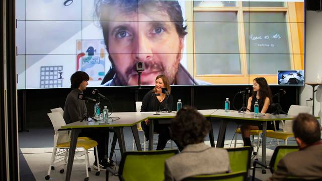 Tres personas participan en un panel de discusión mientras otra persona aparece hablando a través de una videoconferencia en una pantalla grande al fondo.