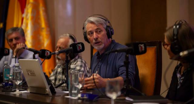 Un grupo de personas participa en una discusión en un estudio de radio con micrófonos y botellas de agua sobre la mesa.