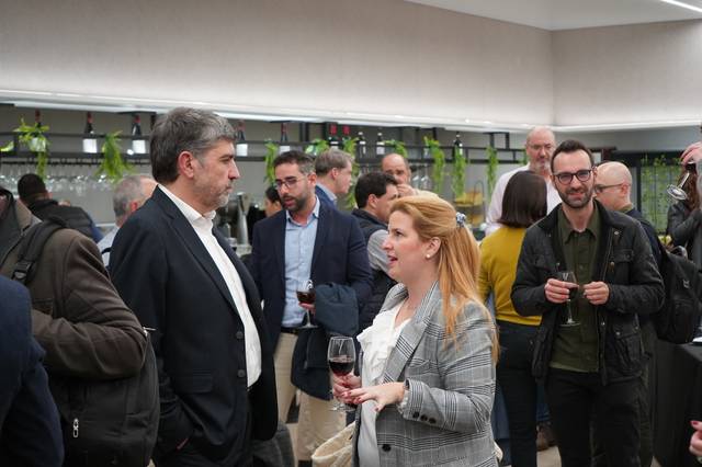 Una mujer y un hombre conversan mientras sostienen copas de vino en un evento social con más personas al fondo.