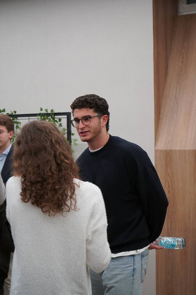 Un hombre con gafas y suéter negro charlando con una mujer de cabello largo en un entorno interior.