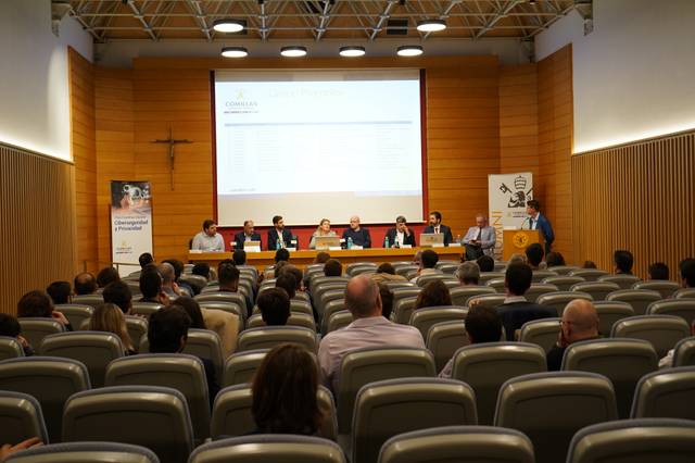 Personas en una sala de conferencias durante una presentación, con un panel de ponentes al frente y una audiencia sentada.