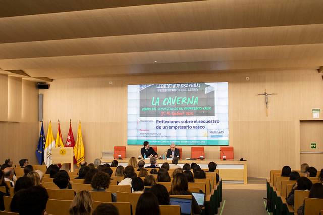 Una conferencia en un auditorio con dos ponentes y una pantalla grande mostrando una presentación titulada 'La Caverna'.