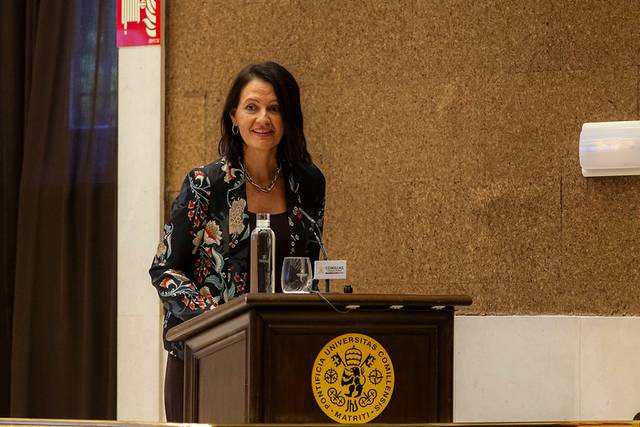 Una mujer sonriente está de pie detrás de un podio en un salón de conferencias con un emblema universitario.