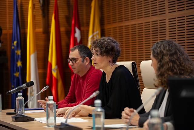 Tres personas sentadas en una mesa durante una conferencia, con micrófonos y botellas de agua delante de ellas.