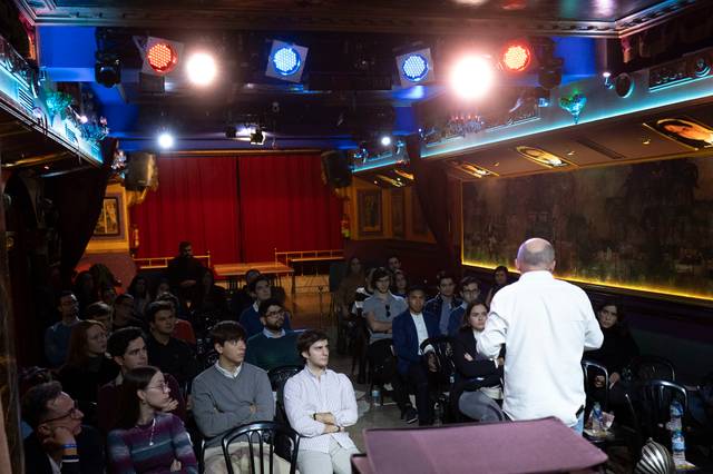 Un hombre habla frente a un grupo de personas sentadas en un teatro iluminado con luces de colores.