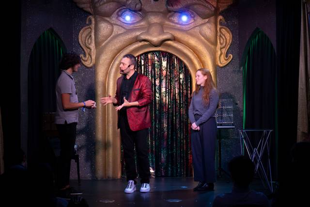 Tres personas en un escenario decorado con una cara gigante, hablando frente a un público.