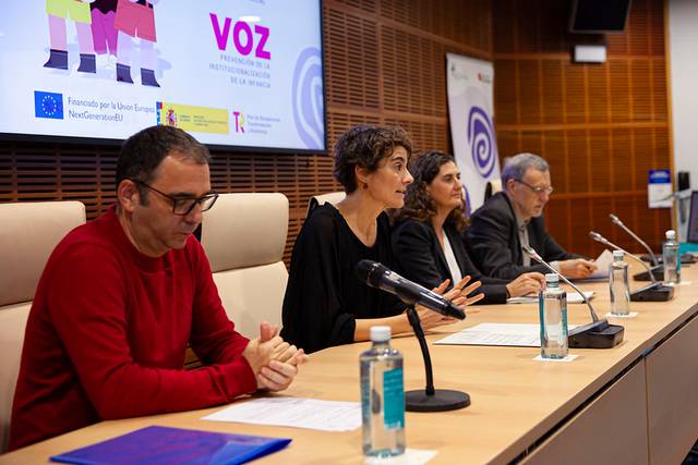 Cuatro personas sentadas en una mesa durante una conferencia, con micrófonos y botellas de agua delante de ellos, en una sala con un cartel al fondo.