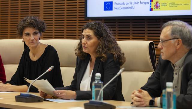 Tres personas sentadas en una mesa durante una reunión, con una mujer hablando en el centro y un cartel de la Unión Europea al fondo.