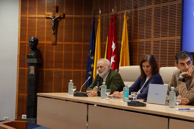 Tres personas sentadas en una mesa en un panel de discusión con banderas de España y la Unión Europea en el fondo.