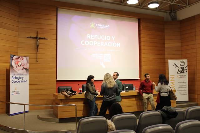 Varias personas conversan en una sala de conferencias con un proyector mostrando la temática 'Refugio y Cooperación'.