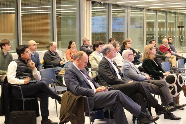 Un grupo de personas sentadas mirando en dirección a un evento o presentación en una sala amplia y moderna.