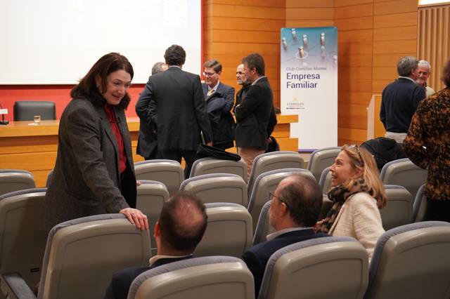 Una mujer sonriente se encuentra hablando con otras personas en un auditorio con asientos vacíos y algunas personas de pie al fondo.