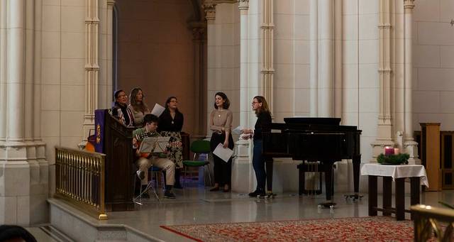 Grupo de personas realizando una presentación musical dentro de una iglesia, con un piano y partituras visibles.