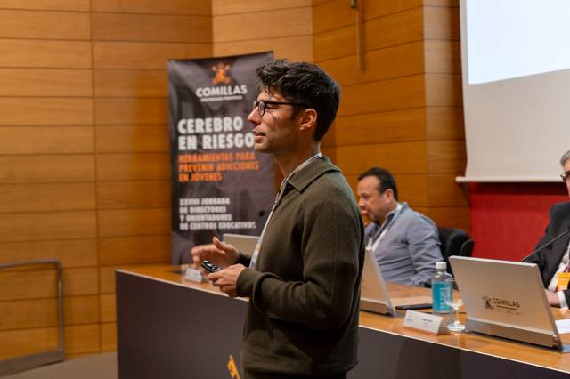 Un hombre de pie dando una presentación en una conferencia, con un cartel de Comillas sobre 'Cerebro en Riesgo' al fondo.