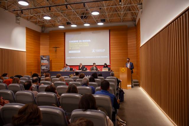 Una persona está dando un discurso en un podio mientras otras personas se sientan en un panel de discusión en un auditorio.