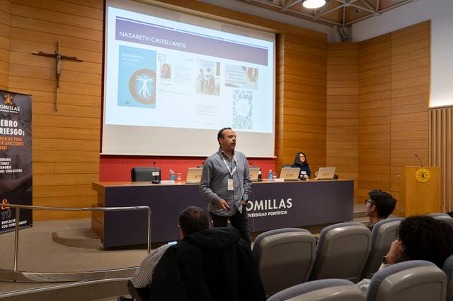 Un hombre está presentando en un auditorio universitario frente a una audiencia y una pantalla que muestra una presentación de PowerPoint.