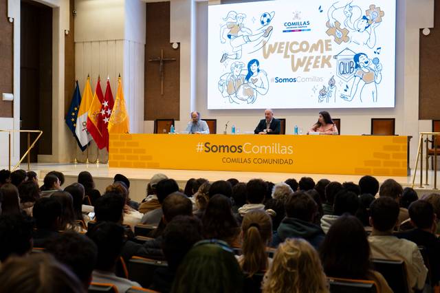 Tres ponentes están sentados frente a una audiencia de estudiantes durante un evento de bienvenida en una universidad, con banderas y una gran ilustración en la pared.