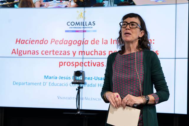 Una mujer está hablando en un evento sobre pedagogía, de pie frente a una pantalla con texto y logos universitarios.