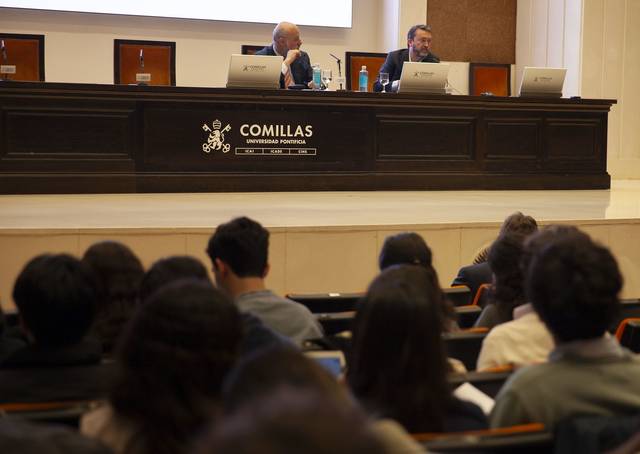 Dos personas están presentando en un evento académico ante una audiencia en la Universidad Pontificia Comillas.