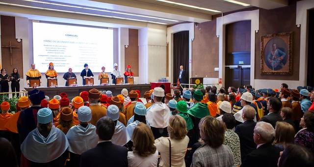 Una ceremonia de graduación en un auditorio con personas usando togas y birretes de colores.