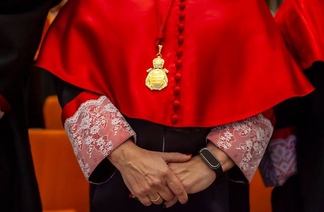 Primer plano de una persona vestida con toga roja y detalles de encaje, llevando un medallón dorado y un reloj moderno.