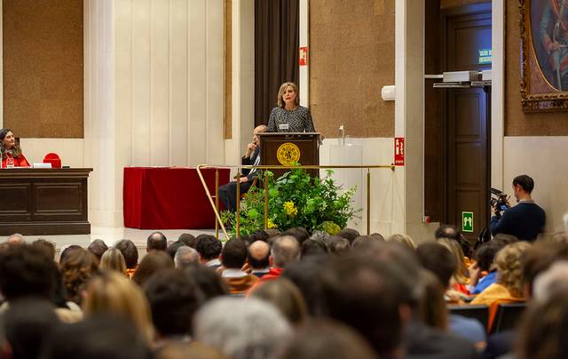 Una mujer está dando un discurso desde un podio frente a una audiencia numerosa en un salón grande y elegante.