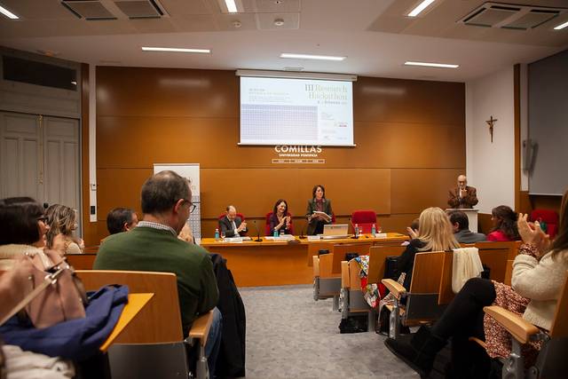 Una conferencia con un grupo de personas sentadas en un auditorio, mientras una mujer habla en el podio.