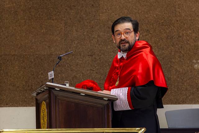 Un hombre con toga y birrete habla desde un podio en un evento académico.