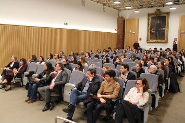 Una audiencia sentada en un auditorio con un fondo de paneles de madera y una pintura en la pared.