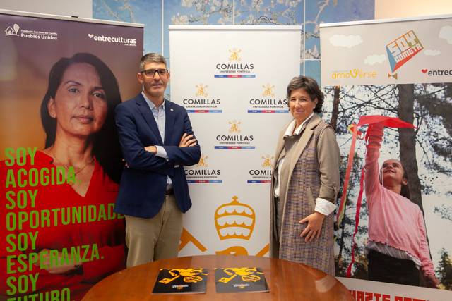 Una imagen que muestra a dos personas de pie, con carteles de fondo y una mesa con objetos sobre ella.