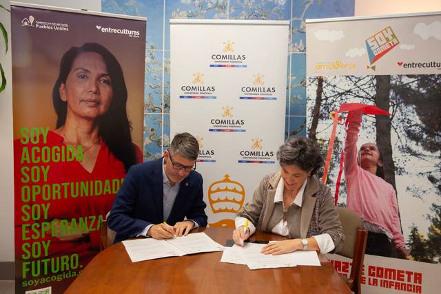 Dos personas firman documentos en una mesa, rodeados de carteles y banderas que representan diferentes iniciativas y organizaciones.