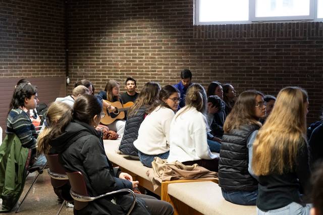 Un grupo de jóvenes sentados en un salón mientras uno de ellos toca la guitarra.