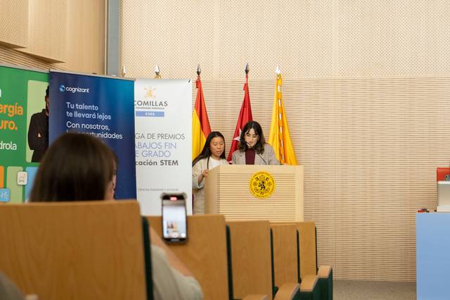 Dos estudiantes presentan un proyecto en un evento académico mientras son observadas por una audiencia.