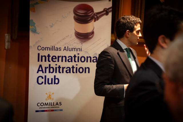 La imagen muestra a un hombre de pie junto a un cartel que promociona el Club de Arbitraje Internacional de Alumni de Comillas.