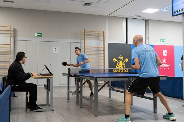 Dos jugadores están compitiendo en una partida de tenis de mesa mientras un árbitro observa desde una mesa.