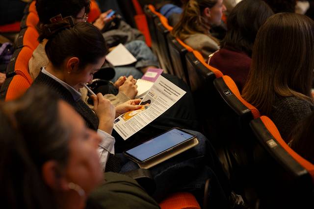 Una mujer está sentada en una sala, enfocada en un documento que sostiene mientras otros asistentes están en el fondo.