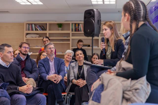 Una mujer habla en un panel frente a un público atento en un ambiente de reunión.