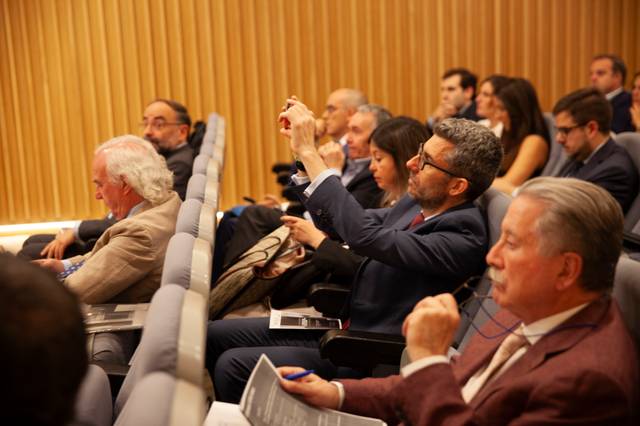 Un grupo de personas sentadas en un auditorio durante una conferencia, algunas de ellas tomando notas o usando sus teléfonos móviles.