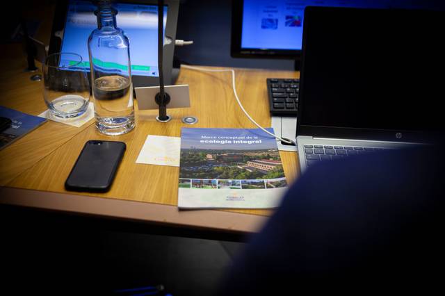 Una mesa de trabajo con una laptop, un teléfono móvil, una botella de agua y un folleto sobre ecología.