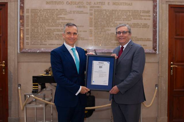 Dos hombres sonríen mientras sostienen un marco con un certificado en un ambiente formal.
