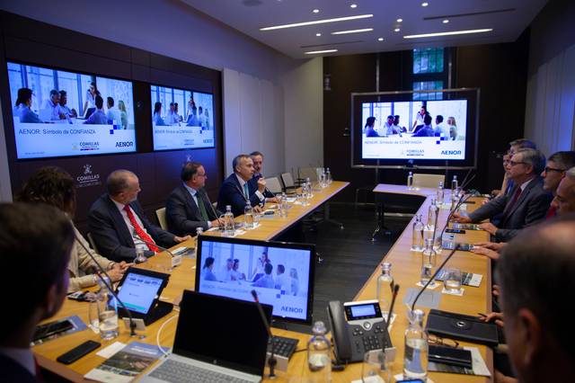 Una reunión empresarial en una sala de conferencias con varias personas sentadas alrededor de una mesa y pantallas digitales mostrando contenido.