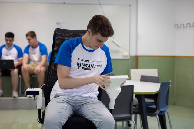 Un joven sentado en una silla, interactuando con un dispositivo, mientras otros estudiantes lo observan en un aula.
