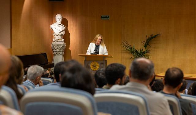 Una presentadora habla frente a un público en un auditorio con un busto de mármol detrás.