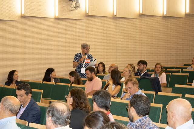 Una reunión en un auditorio con varias personas sentadas y una mujer de pie hablando al micrófono.